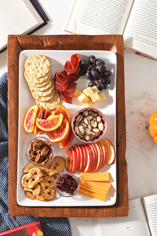A white platter on a wooden tray serves as a snack board with sliced apples, oranges, cheeses, pretzels, grapes, pepperoni and more