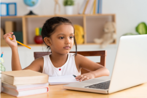 a young child is sitting at a desk with two books to her left (which are closed) and a notebook in front of her with a pencil in her hand which is raised in question. Directly in front of her is an open laptop.