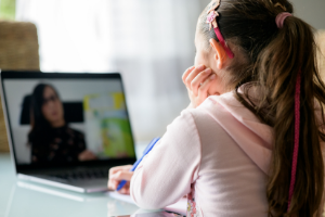 young child sitting at a desk, staring at an open laptop which shows a woman, presumably a teacher, in the middle of a lesson.