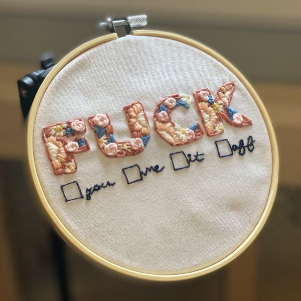 A hoop embroidery. The word "fuck" is sewn in floral, capital letters. Below, in simple black lines, is a horizontal checklist from which one can choose "you," "me," "it," and "off."