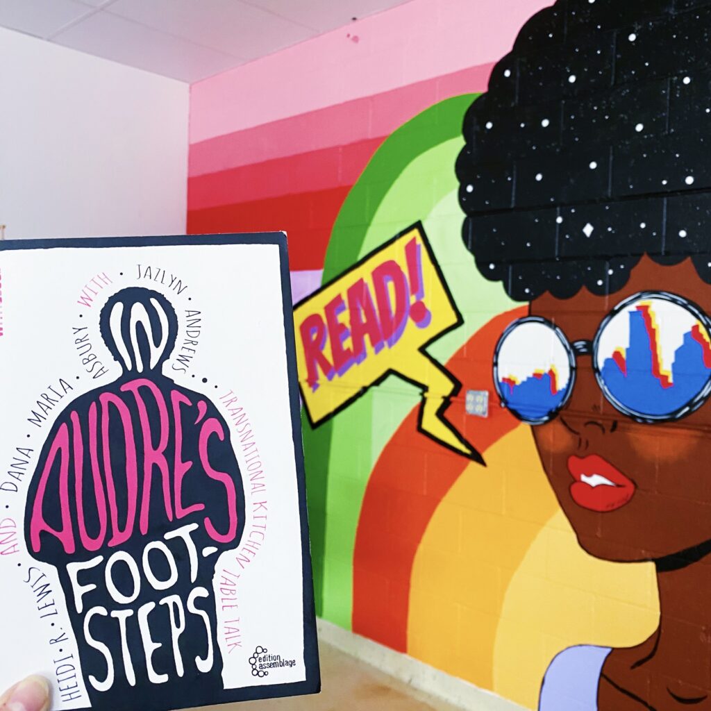 Book cover of In Audre's Footsteps in front of a mural featuring a Black woman with a speech bubble that says READ!