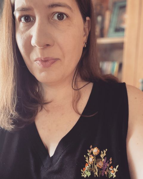 Photograph of me (a white woman with long, brown hair in her 40s) in a black tank top, with flowers embroidered over my heart.
