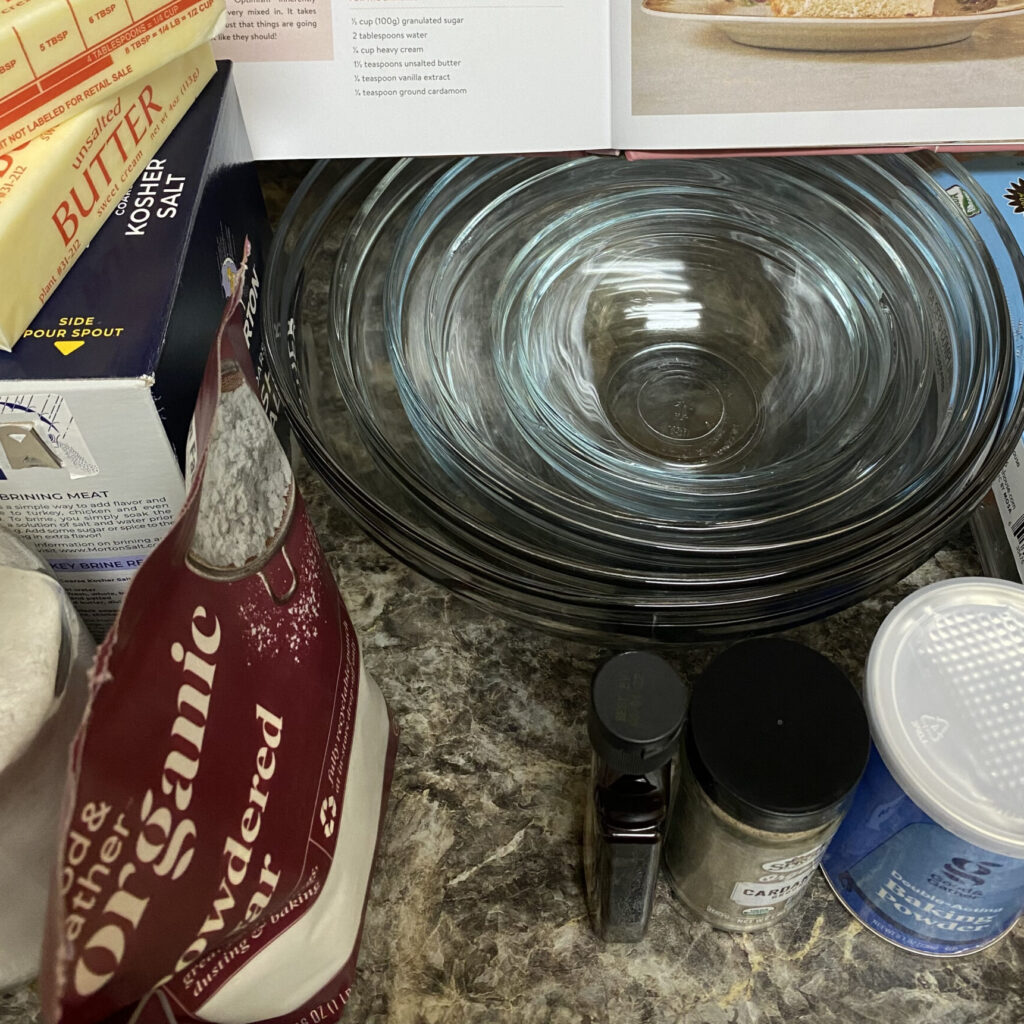Baking by Feel cookbook atop a set of glass bowls surrounded by various baking ingredients (eggs, baking powder, cardamom, vanilla extract, variety of baking ingredients (eggs, baking powder, cardamom, vanilla extract, powdered sugar, kosher salt, butter)