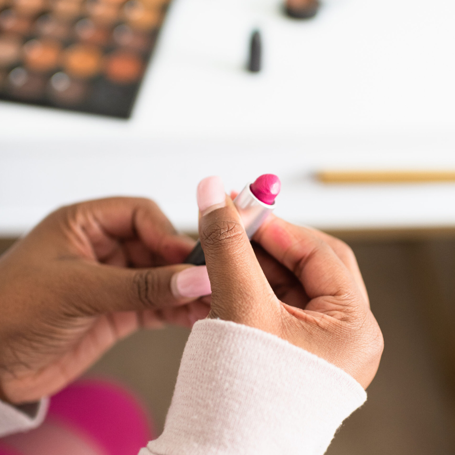 How Our Performance of Beauty and Womanhood Is a Lifelong Trap - closeup on the hands of a Black, feminine-presenting person applying lipstick, with a makeup palette in the background