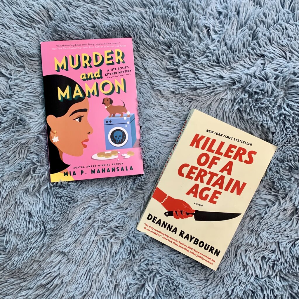 books sit on a fuzzy blue blanket. Books include Murder and Mamon by Mia P. Manasala and Killers of a Certain Age by Deanna Raybourn